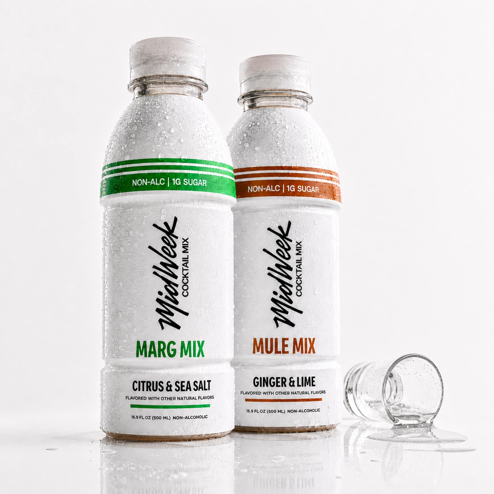 Two Midweek Cocktail Mix bottles, one Citrus & Sea Salt (Marg Mix) with a green band and one Ginger & Lime (Mule Mix) with an orange band, both covered in condensation, with an empty shot glass on a white reflective surface.
