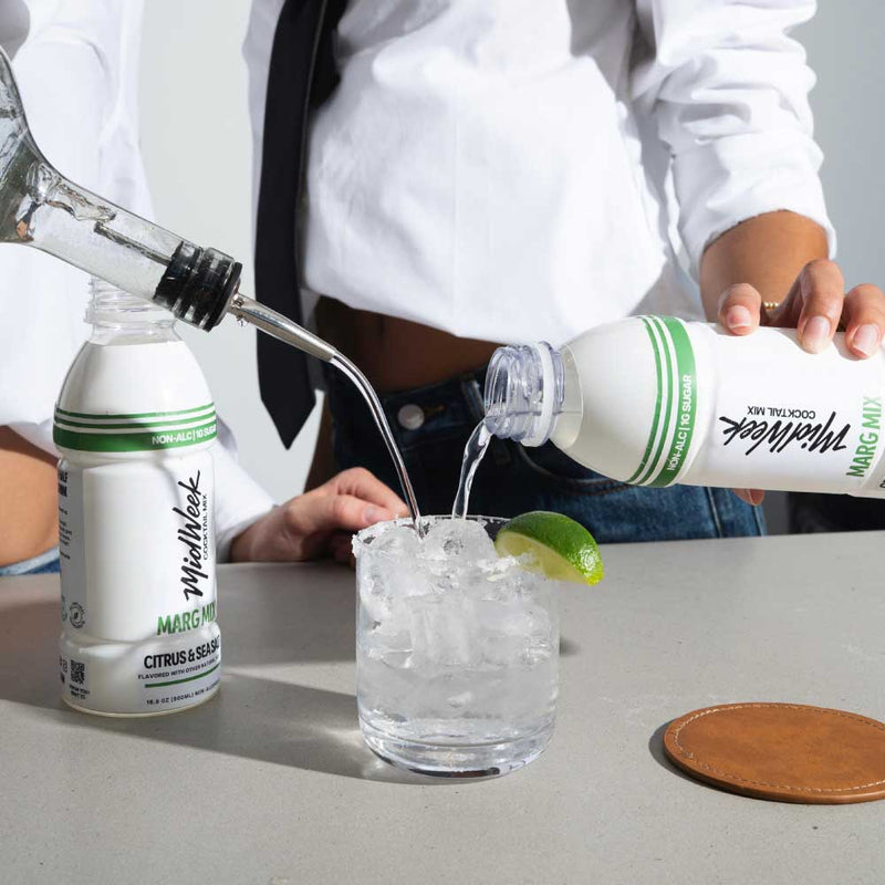 Person pouring from a Midweek Mixers bottle into a glass with ice on a table.